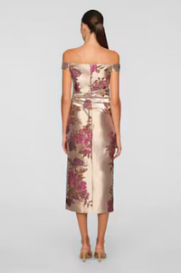 A woman is seen from the back wearing an elegant, off-the-shoulder tea-length dress with floral patterns in shades of pink and gold.