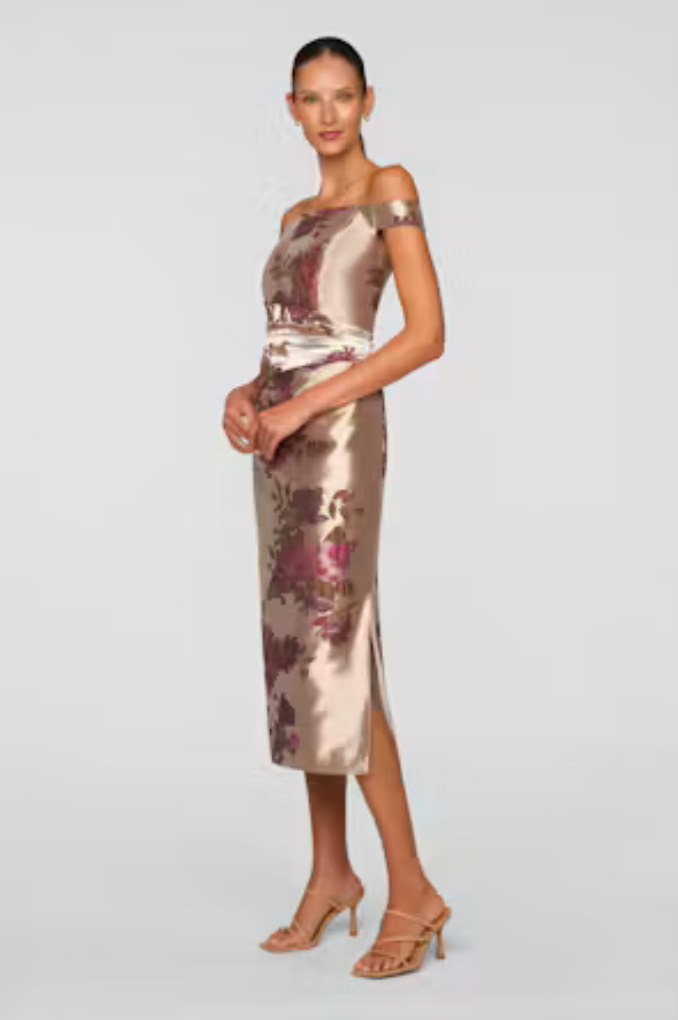 A model wears a sleek, off-the-shoulder tea-length dress in a shimmering beige fabric with floral patterns.