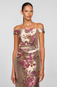 A model showcases an elegant off-the-shoulder gown adorned with a floral pattern in shades of pink and gold, perfect for formal occasions like a gala or wedding. This stunning dress is available near Apex, Cary, and Raleigh.