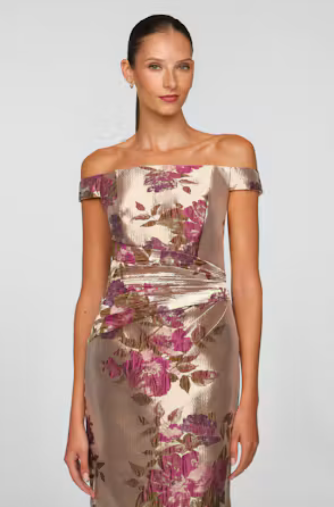 A model showcases an elegant off-the-shoulder gown adorned with a floral pattern in shades of pink and gold, perfect for formal occasions like a gala or wedding. This stunning dress is available near Apex, Cary, and Raleigh.