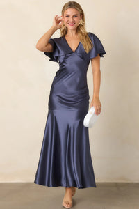 A woman is wearing a steel blue satin maxi dress featuring flutter sleeves and a plunging V-neckline. This elegant gown is perfect for formal events like a black tie wedding or gala, and is conveniently located near Apex, Cary, and Raleigh.