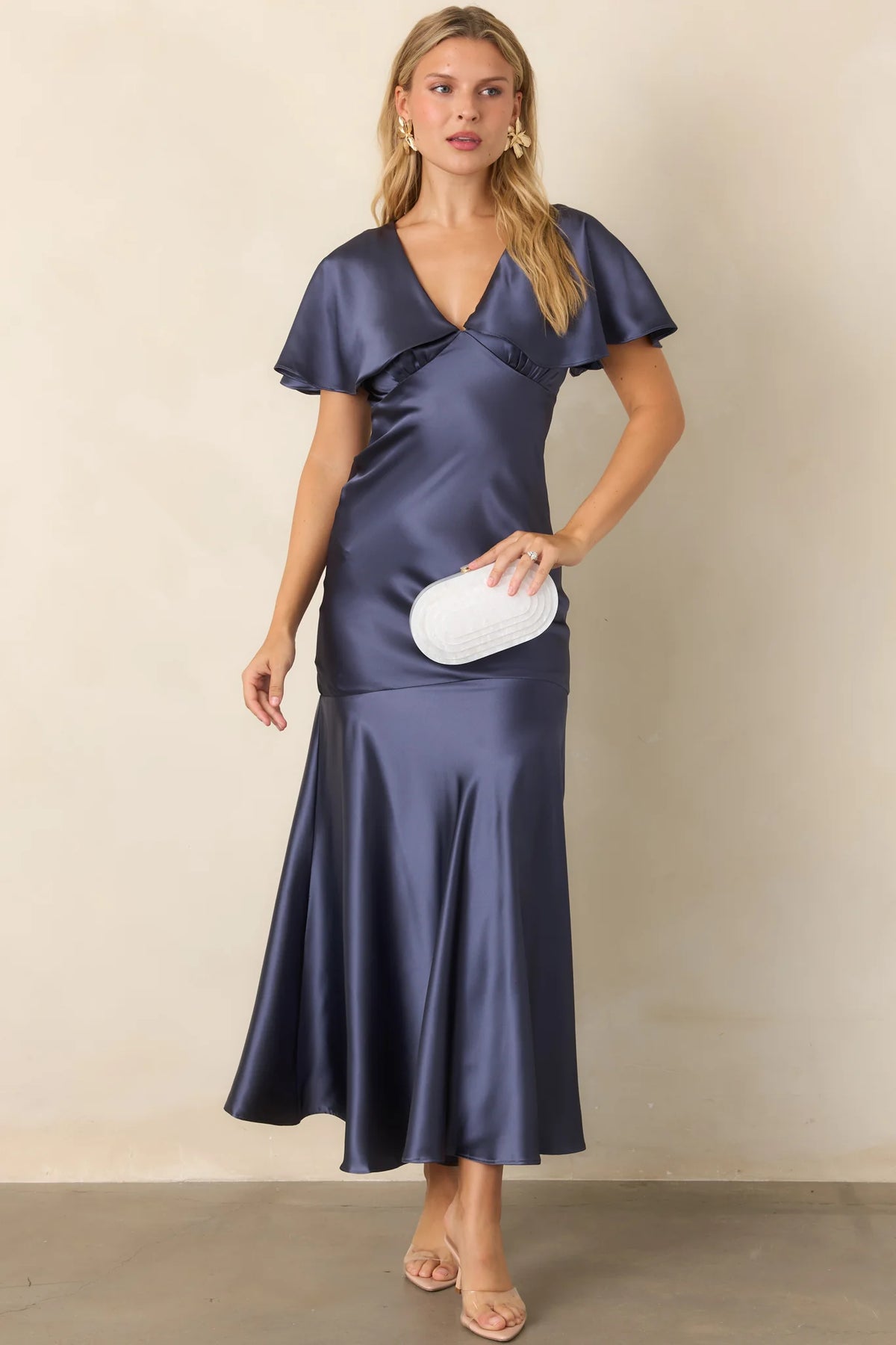 A model showcases a steel blue satin maxi dress with flutter sleeves and a plunging V-neckline, perfect for elegant occasions like galas or black tie weddings. This stunning gown is conveniently located near Apex, Cary, and Raleigh.