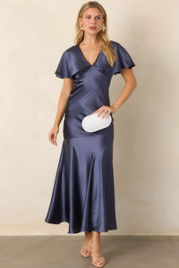 A model showcases a steel blue satin maxi dress with flutter sleeves and a plunging V-neckline, perfect for elegant occasions like galas or black tie weddings. This stunning gown is conveniently located near Apex, Cary, and Raleigh.