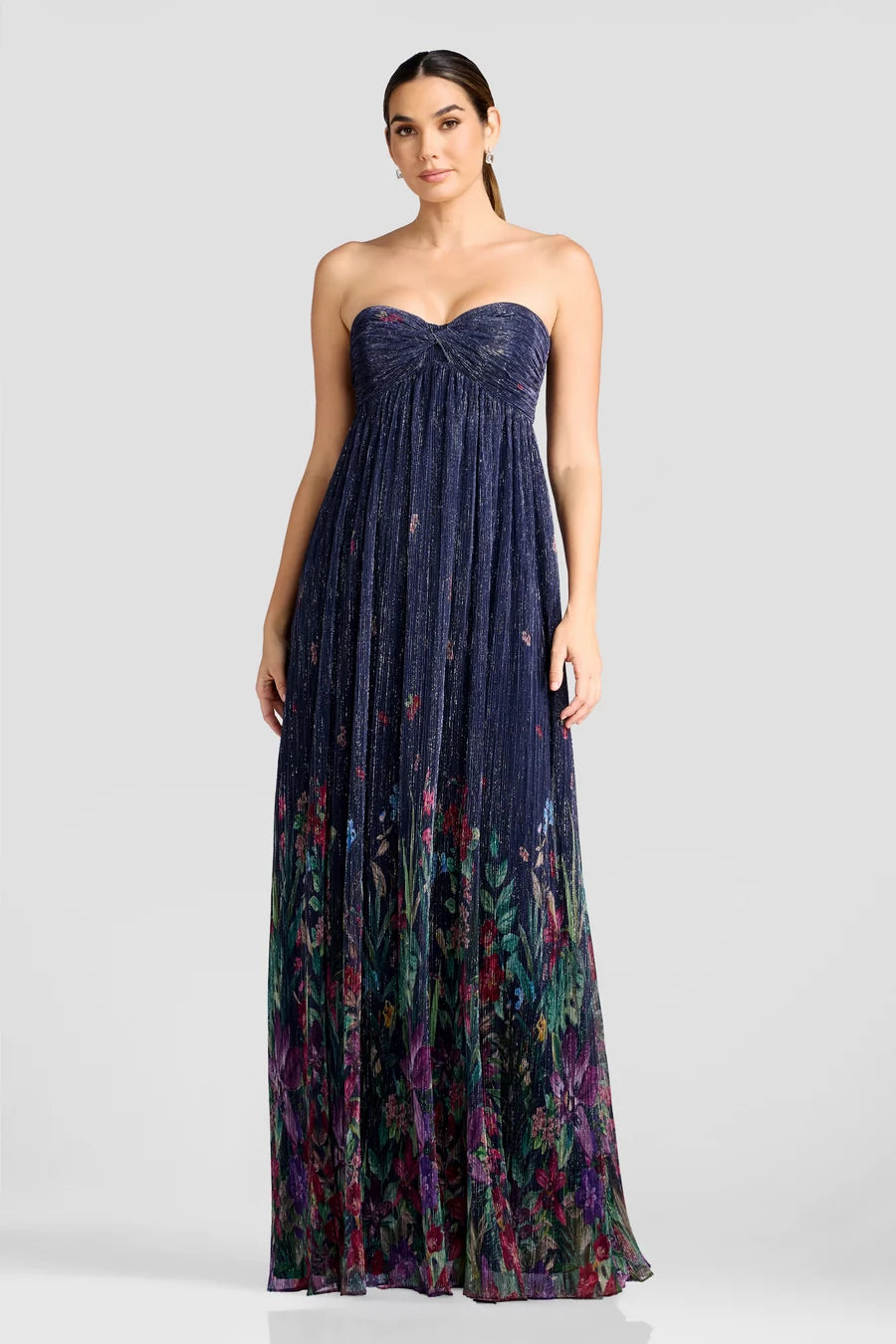 A model showcases a stunning strapless floor-length gown featuring shimmering pleated fabric with a floral design at the hem.