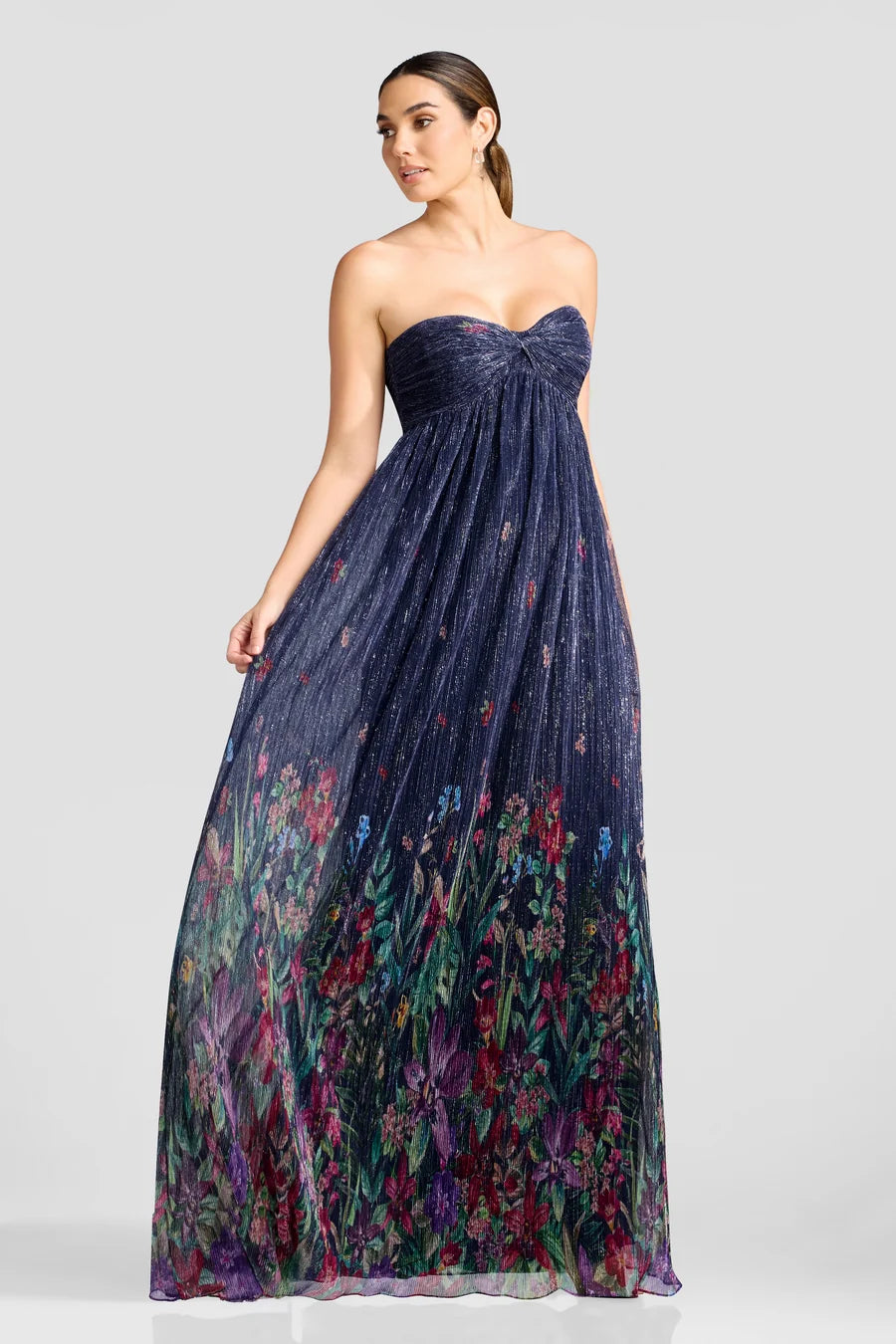 A woman models a stunning strapless gown with a pleated navy bodice and a flowing floral print skirt in vibrant colors, perfect for occasions like a black-tie wedding or gala.