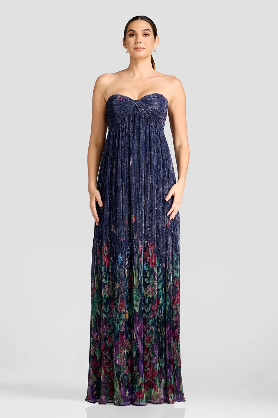 A model showcases a stunning floor-length gown featuring a strapless design with a pleated bodice and a vibrant floral pattern at the hem.