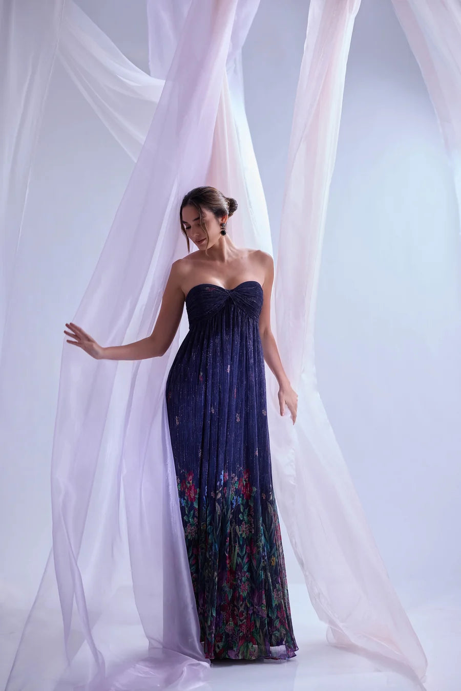 A model stands gracefully in a strapless, floor-length gown featuring a shimmering navy pleated fabric with floral accents at the hem.