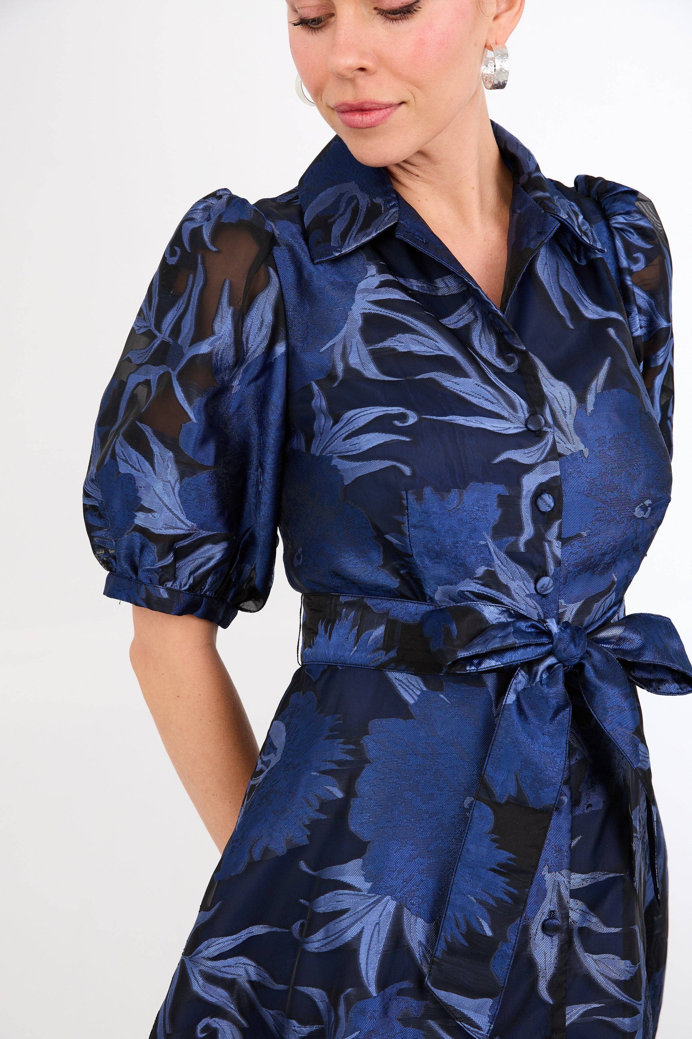 A woman models a stylish navy blue gown featuring a floral design in shades of royal blue, with puffed sleeves and a tie waist.