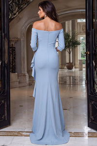 A woman stands gracefully in a light blue off-the-shoulder gown with a fitted silhouette and a cascading ruffle at the back, set against an elegant foyer with marble flooring and large windows.
