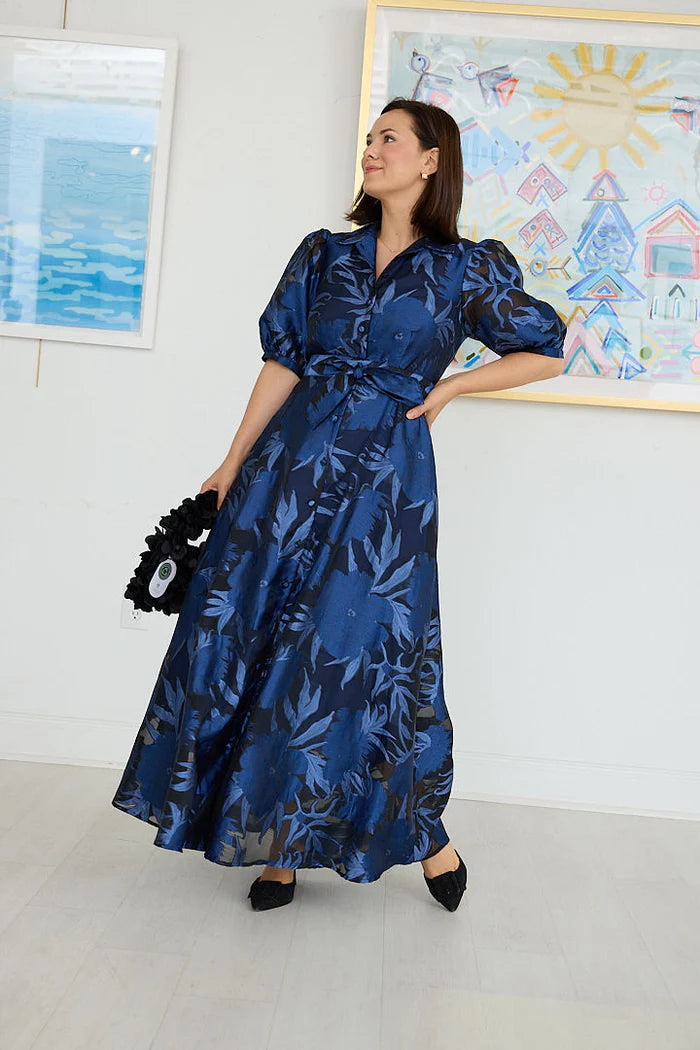 A woman poses confidently in a long, flowing gown featuring a royal blue floral pattern and puffed sleeves.