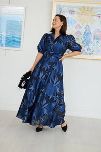 A woman poses confidently in a long, flowing gown featuring a royal blue floral pattern and puffed sleeves.