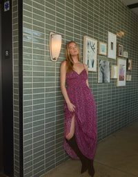 A model poses in a vibrant, floral smocked dress with a thigh-high slit, standing against a textured green tiled wall adorned with various framed artworks.