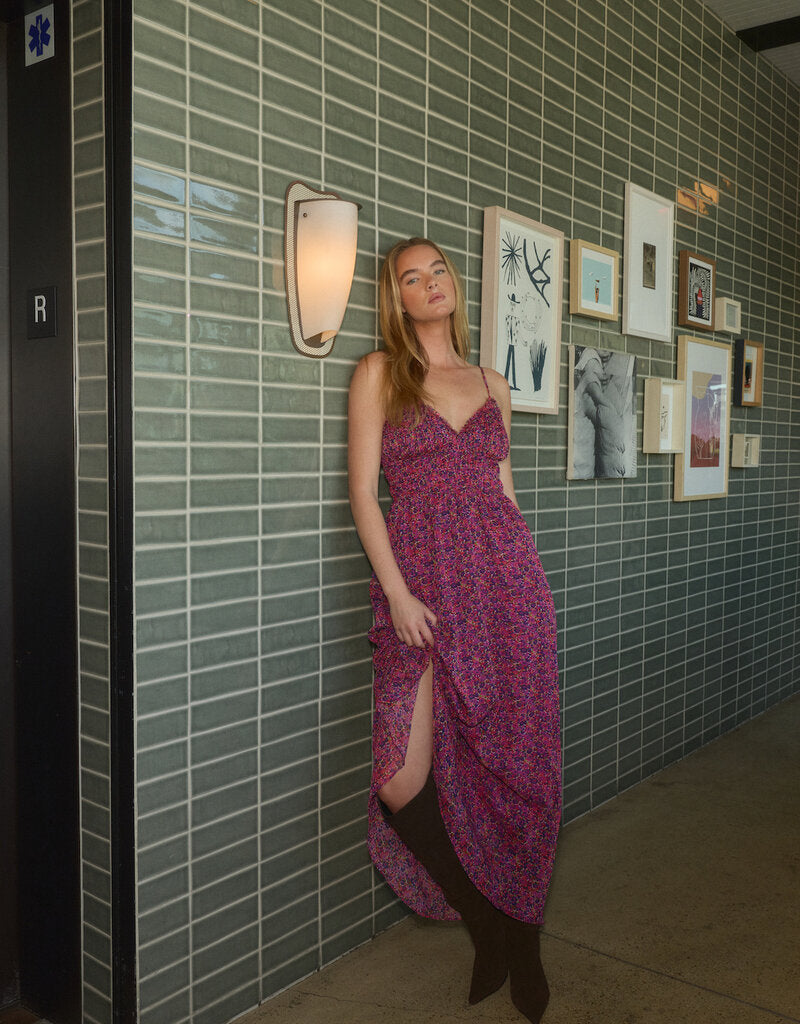A model poses in a vibrant, floral smocked dress with a thigh-high slit, standing against a textured green tiled wall adorned with various framed artworks.