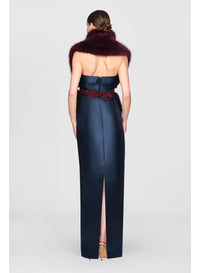 A model is shown from the back wearing a sleek, navy gown with a thigh-high slit and a burgundy bow accent at the waist.