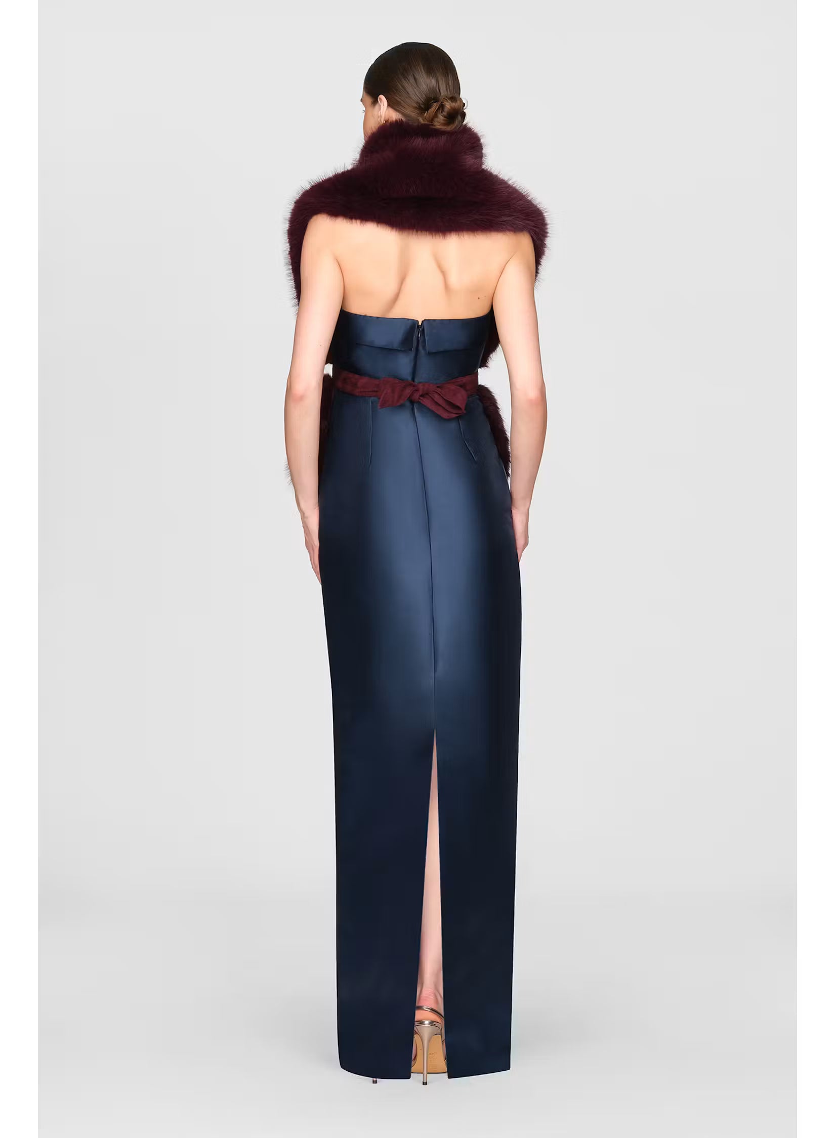 A model is shown from the back wearing a sleek, navy gown with a thigh-high slit and a burgundy bow accent at the waist.